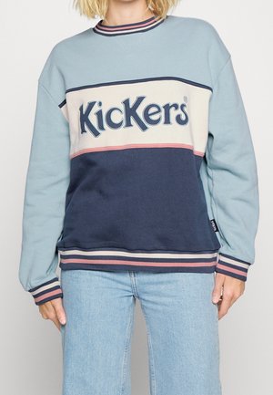 Femme portant un sweat Kickers bicolore bleu, crème et rose avec un jean bleu sur un fond uni.