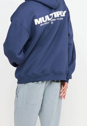 Personne portant un sweat-shirt bleu marine avec le texte « MULTIPLY » dans le dos et un pantalon gris clair avec de grandes poches, main reposant sur le côté.