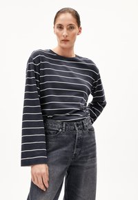 Donna con i capelli raccolti che indossa una maglietta a righe blu scuro e bianca con maniche lunghe, infilata in un paio di jeans neri sbiaditi a vita alta, in piedi con una mano sulla vita.