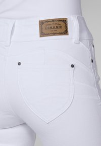 Balti džinsi ar detalizētu aizmugurējo kabatu ar sirds formas dizainu, smalku šuvumu un ādas marķējumu ar zīmola informāciju.