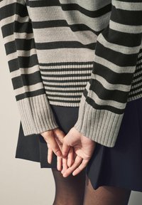 Pull rayé gris et noir, avec poignets et ourlet côtelés. Le mannequin porte une jupe noire. Mains jointes dans le dos.