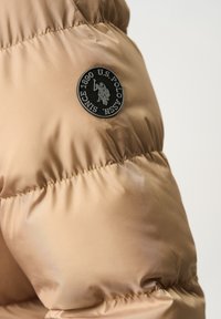 Béžová puffová bunda s prešívanou textúrou a okrúhlym čiernym logom označeným "U.S. Polo ASSN. Since 1890" na pleci.