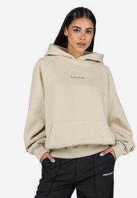Beige oversized hoodie with a front pocket and "PEGADOR" logo in black. Features a drawstring hood and ribbed cuffs. Paired with black pants.