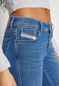 Jeans de mezclilla azul ajustados con un ligero desgaste, cinco bolsillos y una etiqueta de marca en la parte posterior; tela texturizada con detalles de costura visibles.