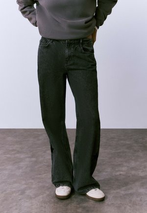Persona que lleva unos pantalones vaqueros holgados negros con patrones de estrellas en los laterales, sudadera gris y zapatillas blancas, de pie sobre un suelo gris.