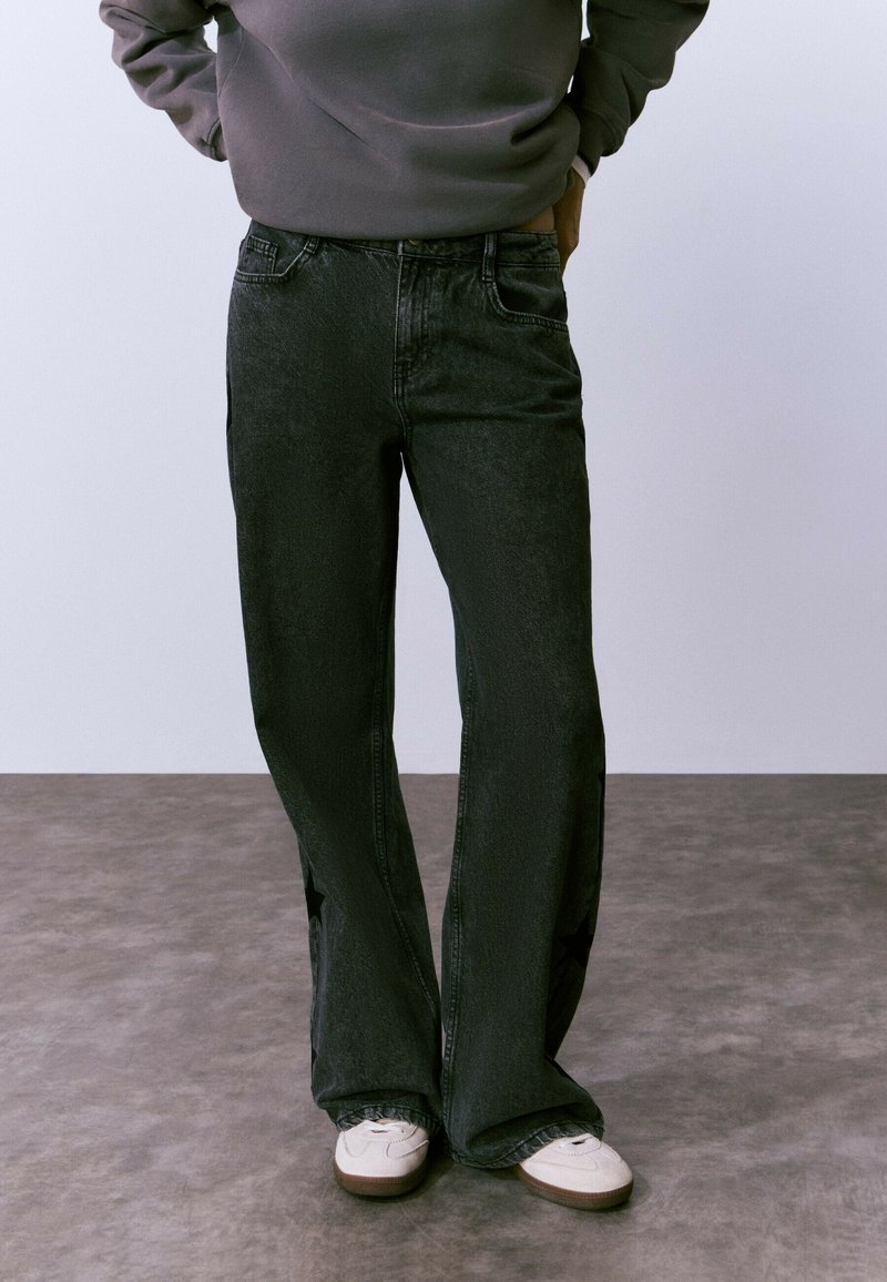 Persona que lleva unos pantalones vaqueros holgados negros con patrones de estrellas en los laterales, sudadera gris y zapatillas blancas, de pie sobre un suelo gris.