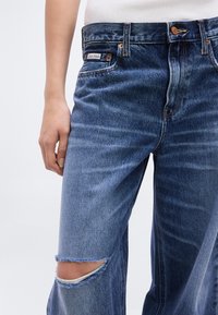 Κοντινό πλάνο ατόμου που φοράει μπλε τζιν με σκίσιμο στο γόνατο, εμφανή ετικέτα Calvin Klein και λευκό πουκάμισο φορεμένο μέσα.