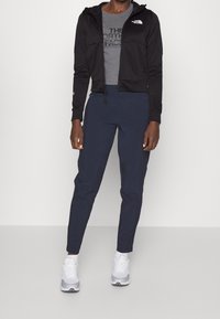 Felpa nera con zip e texture a costine, t-shirt grigia con logo e pantaloni blu tapered. Abbinati a scarpe da ginnastica bianche.