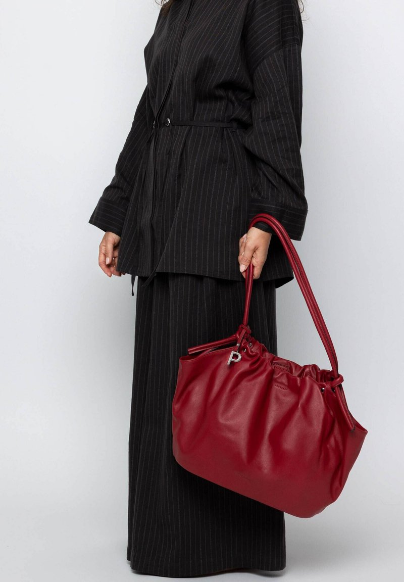 Red leather handbag with a gathered top, adjustable straps, and silver hardware. Worn with a black pinstriped outfit.