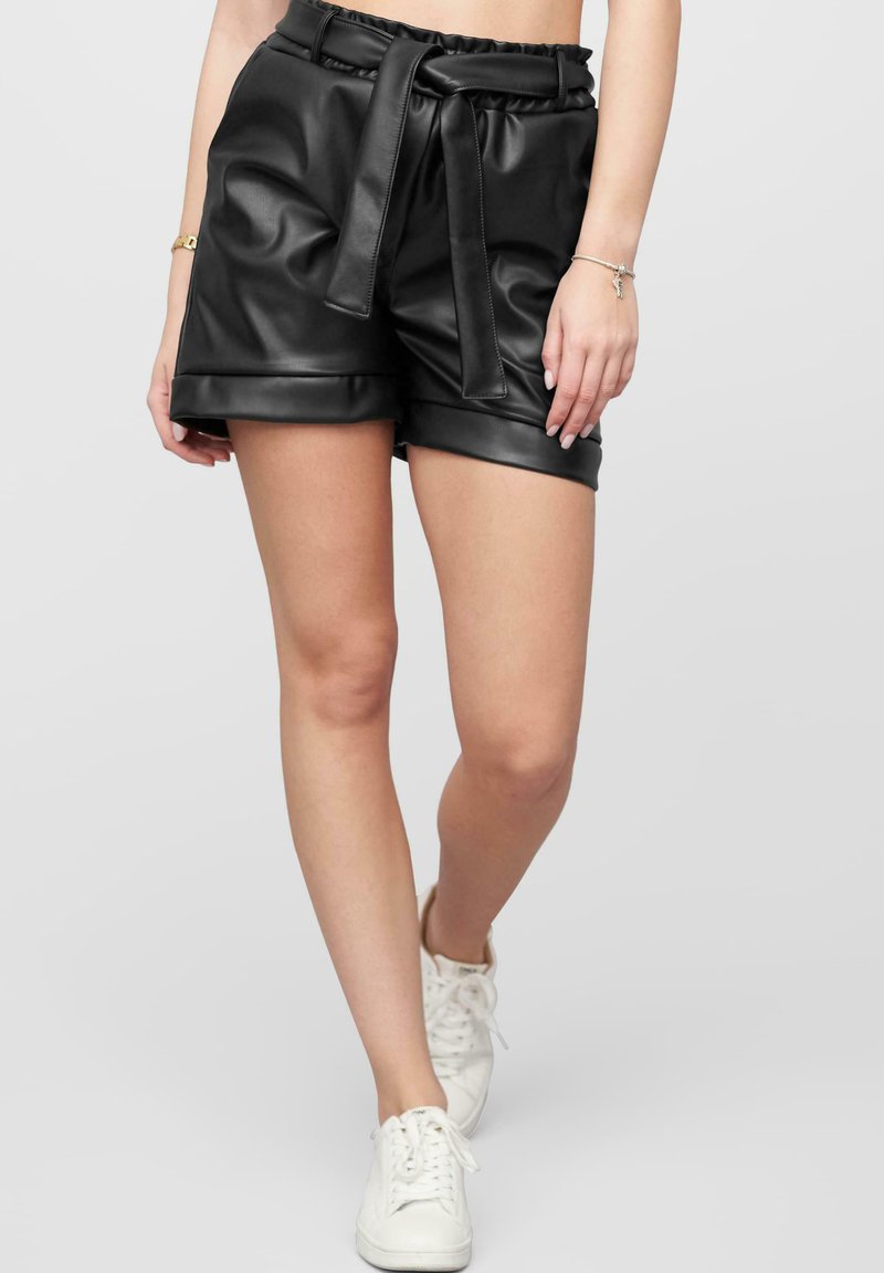 Pantalones cortos de piel sintética negra con una cinturilla con volantes y un cinturón de lazo, que presentan una textura suave y dobladillos levantados. Combinados con zapatillas blancas.