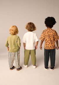 Három gyerek áll háttal egymásnak, zöld, fehér és narancssárga mintás pólókat viselve, bőszárú nadrágokkal, különböző textúrákat és színeket bemutatva.