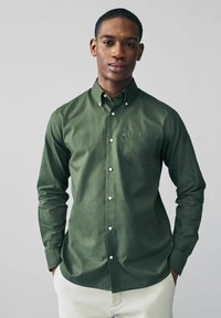 Camisa verde de botones fabricada en tejido suave, con un bolsillo en el pecho y botones blancos. El modelo lleva pantalones de color claro.