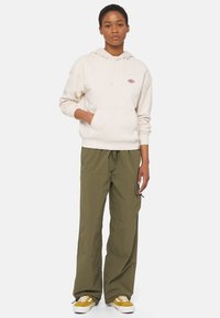 Ljust beige hoodie med en framficka och broderad logotyp, tillsammans med lösa olivgröna cargobyxor och gula och vita sneakers.