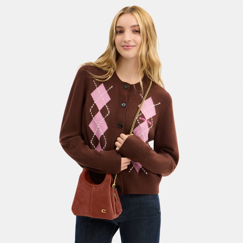 Femme portant un cardigan marron avec un motif losange rose, un jean foncé et un sac à main en daim rouge avec une lanière en chaîne dorée.