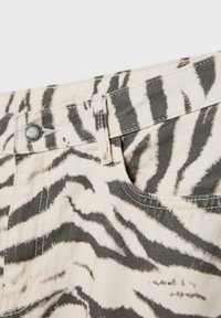 Gros plan sur un tissu beige à rayures zèbre noires, montrant une boucle pour doigt, un bouton en métal et une partie d'une poche avant.