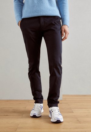 Schwarze Chinos mit schmalem Schnitt, kombiniert mit weißen und lila Sneakers. Das Outfit umfasst einen hellblauen Strickpullover für den Kontrast.