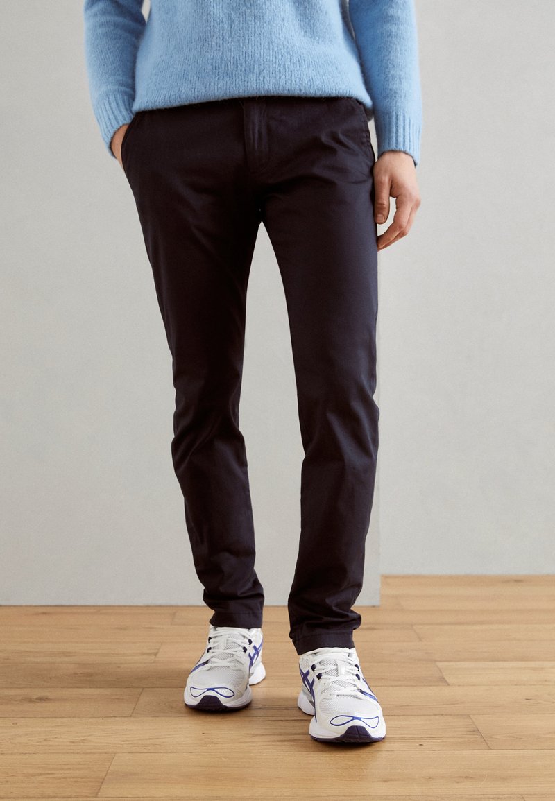 Chino neri con vestibilità affusolata, abbinati a sneakers bianche e viola. L'outfit include un maglione lavorato a maglia blu chiaro per creare contrasto.