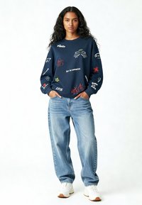 Marineblauwe sweatshirt met verschillende gekleurde grafische prints, comfortabele pasvorm, gecombineerd met losse lichtblauwe jeans en witte sneakers.