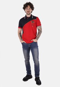 Polo shirt rosso e blu con un design curvo, logo ricamato e dettagli bianchi nel colletto, abbinato a jeans blu e sneakers nere.