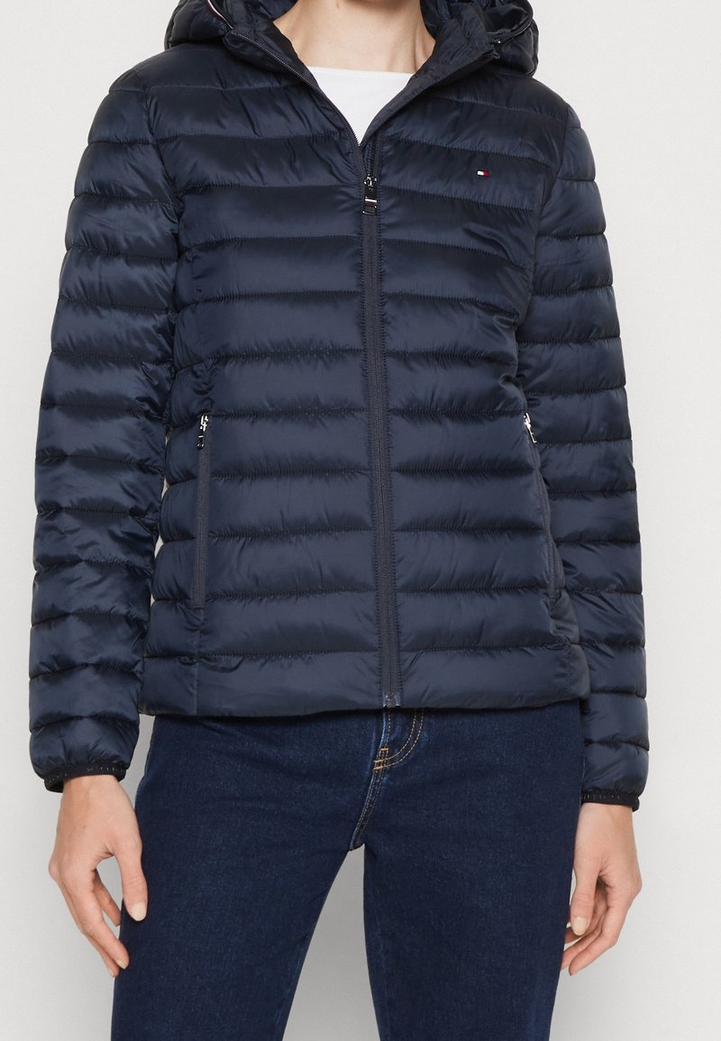 Persona che indossa una giacca a cappuccio trapuntata blu navy, chiusa con la zip, abbinata a jeans blu scuro e una maglietta bianca sotto.