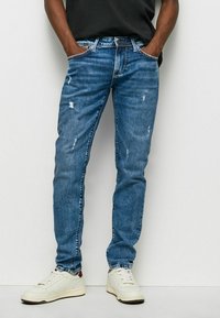 Jeans de mezclilla azul con un ajuste slim, con detalles desgastados y un diseño clásico de cinco bolsillos. Combinados con zapatillas blancas.