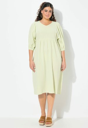 Helles, grünes, knielanges Kleid mit V-Ausschnitt, Puffärmeln und gerafftem Bund. Tragen mit braunen Plateausandalen. Weicher, strukturierter Stoff.