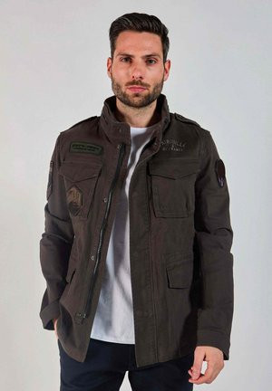 Homme aux cheveux courts et foncés, avec une barbe, portant une veste style militaire vert foncé sur un t-shirt blanc, debout devant un fond clair uni.