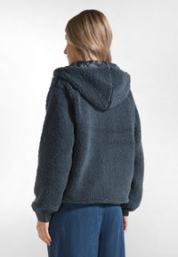 Giacca in pile grigia scura con cappuccio, superficie testurizzata e polsini elasticizzati, abbinata a pantaloni di jeans a gamba larga.