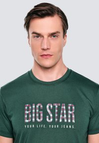 Tumši zaļš kokvilnas T-krekls ar šķērsgriezuma rakstu, kurā ir teksts "BIG STAR" un sauklis "TAVA DZĪVE. TAVI GENE." drukāts zemāk.