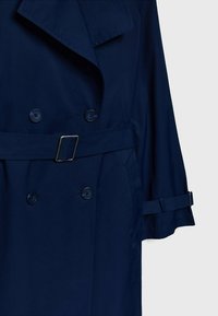 Trench-coat bleu marine croisé avec ceinture et boucle, poignets boutonnés et col à revers sur fond gris clair.