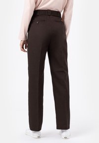 Hnědé kalhoty s vysokým pasem, rovnými nohavicemi a zadními kapsami. Tkanina vypadá texturovaně s hladkým povrchem. Viditelné bílé tenisky.