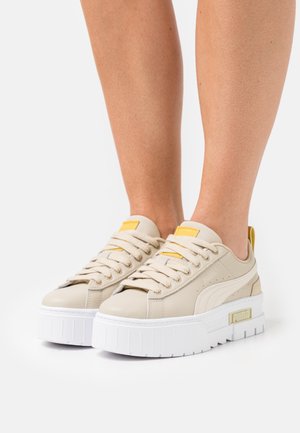 Des baskets plateforme beiges avec de grosses semelles blanches et des accents jaunes, portées sur des jambes nues sur fond blanc.