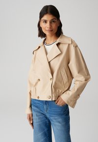 Giacca beige tagliata con design Doppiopetto, grandi bottoni e tasche laterali. Presenta un colletto aperto e una vita elasticizzata.