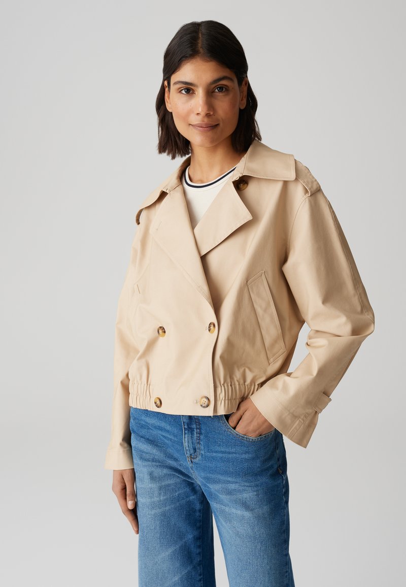 Giacca beige tagliata con design Doppiopetto, grandi bottoni e tasche laterali. Presenta un colletto aperto e una vita elasticizzata.