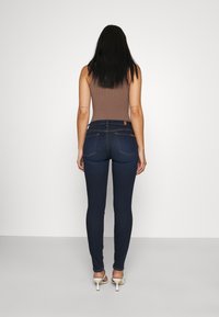 Vaqueros skinny azul oscuro de cintura alta, con detalles de costura sutiles y dos bolsillos traseros, combinados con una blusa sin mangas marrón.