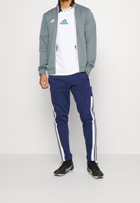 Mand iført hvid Adidas t-shirt, grå zip-up jakke, marineblå Adidas træningsbukser med hvide striber og sorte fodboldstøvler.