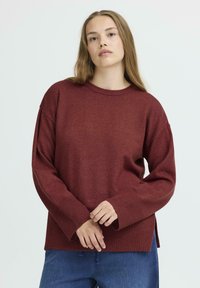 Pull en maille bordeaux avec des manches larges, des poignets côtelés et des fentes sur les côtés. Associé à un jean bleu, créant une silhouette décontractée.