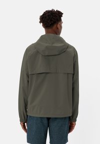 Khaki-grüne, wasserdichte Jacke mit Kapuze, elastischen Bündchen, einem Rückeneinsatz und einer glatten Textur. Kombiniert mit dunklen Shorts.
