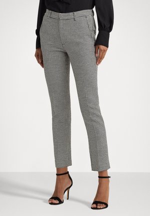 Vrouw draagt zwart-witte slanke broek tot op de enkel met pied-de-poulemotief, een zwart shirt met lange mouwen en zwarte hoge hakken met enkelbandje.