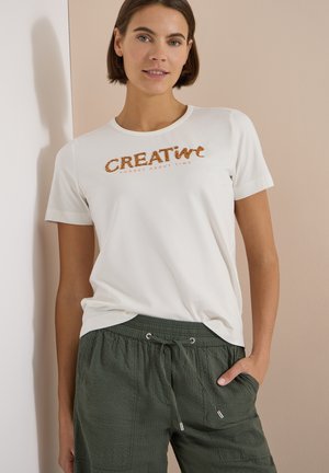 Kvinde iført en hvid t-shirt med teksten "CREATIVE" og grønne bukser med snøre, stående mod en beige væg med den ene hånd i lommen.
