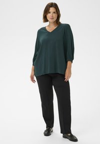 Femme portant un blouse à col en V vert foncé avec des manches 3/4, un pantalon noir droit et des mocassins noirs, se tenant contre un fond blanc uni.
