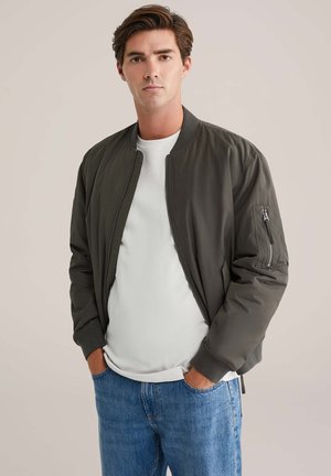 Chaqueta bomber verde oliva con un diseño liso, puños acanalados y un bolsillo con cremallera en la manga izquierda. Lleva puesta una camiseta blanca.