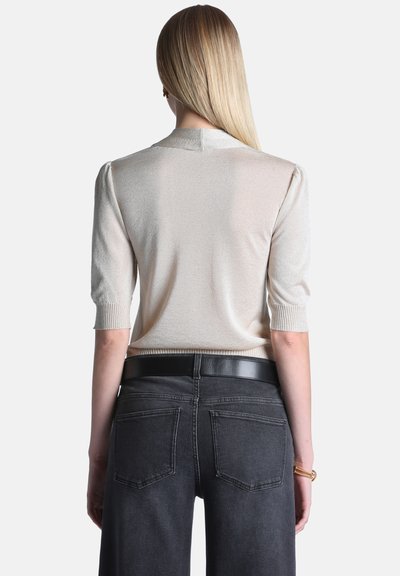 Femme aux longs cheveux blonds portant un haut beige clair à manches courtes et un jean gris foncé avec une ceinture noire, vue de dos.