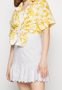 Mini-jupe blanche en broderie anglaise avec un ourlet froncé, associée à une chemise à fleurs jaune à manches courtes nouée à la taille.