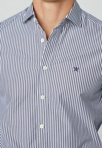 Primo piano di una persona che indossa una camicia a righe verticali blu navy e bianche con un piccolo logo di spade incrociate sul petto.