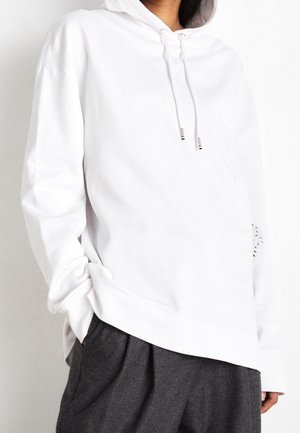 Osoba w białej bluzie z kapturem i sznurkami oraz w luźnych ciemnoszarych spodniach, z rękami częściowo w kieszeniach, na jednolitym tle.