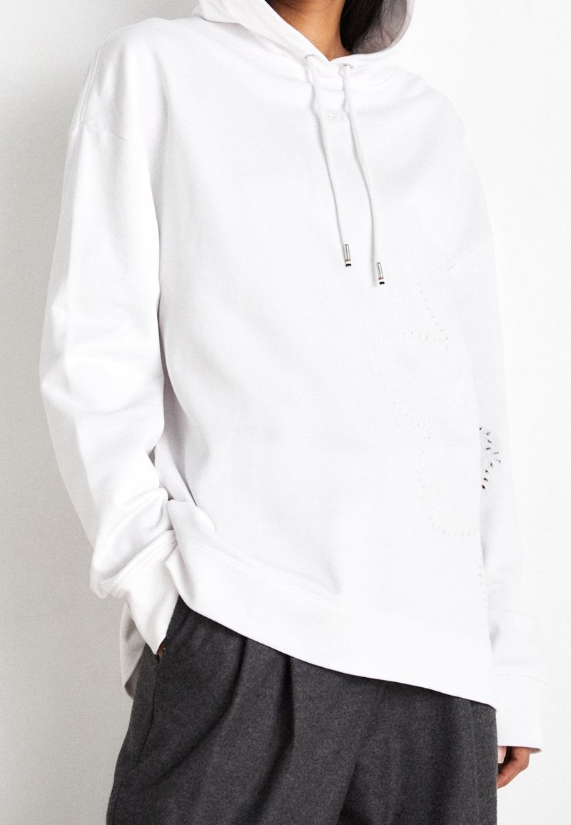 Personne portant un sweat à capuche blanc avec cordons et un pantalon ample gris foncé, mains partiellement dans les poches, sur un fond uni.