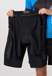 Personne en tenue de cyclisme tenant un cuissard noir Swedemount avec doublure intérieure rembourrée et logo visible sur la ceinture et les jambes.