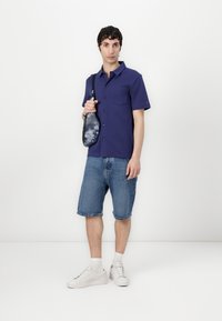 Krótki, fioletowy t-shirt z zapięciem na guziki, zestawiony z szortami dżinsowymi do kolan. Modelka trzyma małą, wzorzystą torebkę na ramię. Białe trampki dopełniają całość.