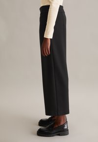 Pantalons larges noirs en tissu lisse, avec une taille haute et une coupe droite, associés à des mocassins noirs.